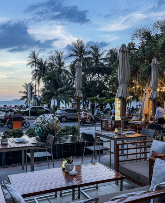 Outdoor beachfront cafe at dusk with palm trees, wooden tables, and diners along a coastal promenade by the sea.