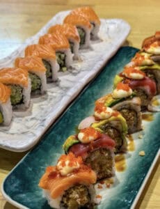 Two long trays of assorted sushi rolls: one with salmon-topped rolls, the other with mixed rolls topped with sauces, avocado, and roe on a teal plate and a white marble plate.