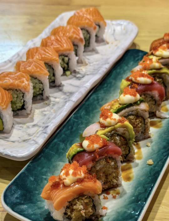 Two long trays of assorted sushi rolls: one with salmon-topped rolls, the other with mixed rolls topped with sauces, avocado, and roe on a teal plate and a white marble plate.