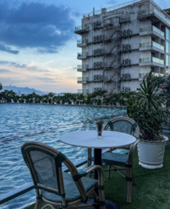 Poolside seating area with a round table and two wicker chairs beside a swimming pool, with apartment building and palm plants in the background.