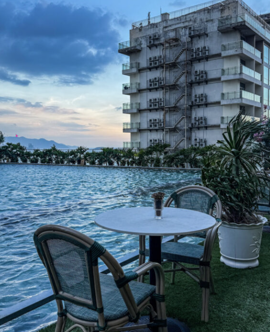 Poolside seating area with a round table and two wicker chairs beside a swimming pool, with apartment building and palm plants in the background.